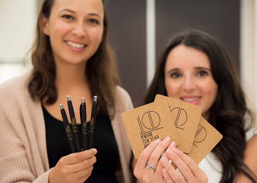 Two NWHM employees holding pens and coasters with the anniversary logo